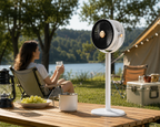 A white pedestal fan stands on a wooden table outdoors at a lakeside campsite, with a woman seated in a folding chair in the background near a tent and trees.