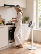 A woman in a bright, modern kitchen stirs a pot on the stove while a white and gray pedestal fan stands on the wooden floor nearby.