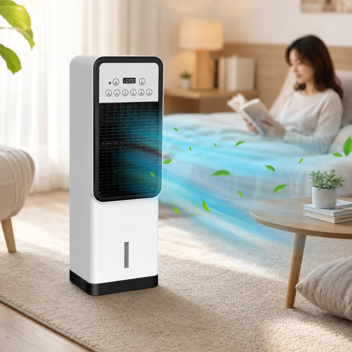 A white and black portable air cooler stands on a beige rug in a bright bedroom. A woman is sitting on a bed reading a book in the background, and blue airflow graphics with green leaves are shown coming out from the front.