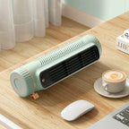 A compact, portable desk fan in a light mint green color with a black front grille, resting on a wooden desk next to a white computer mouse, a cup of latte macchiato, and a laptop.
